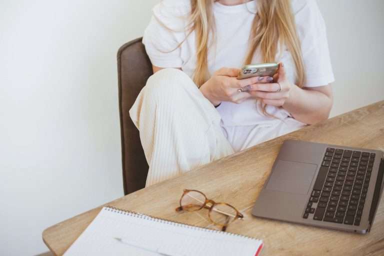 Steuerblog für Selbstständige: Steuerwissen Frau sitzt vor Laptop am Schreibtisch mit Handy in der Hand.