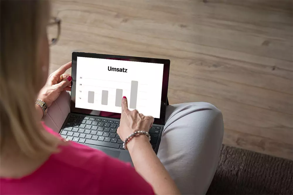 Symbolbild Wechsel Kleinunternehmer zur Regelbesteuerung: Barbara Hülsmann, Steuerberaterin, sitzt am Boden mit Laptop. Der Laptop zeigt einen Umsatzanstieg.